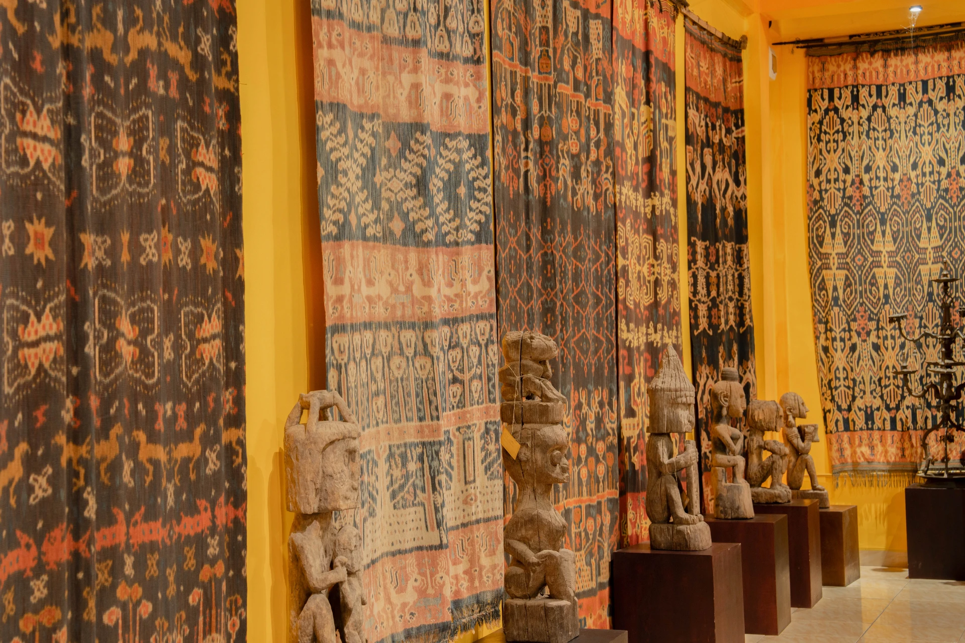 Framed traditional ikat textiles and detailed Balinese wood carvings displayed on red gallery walls.