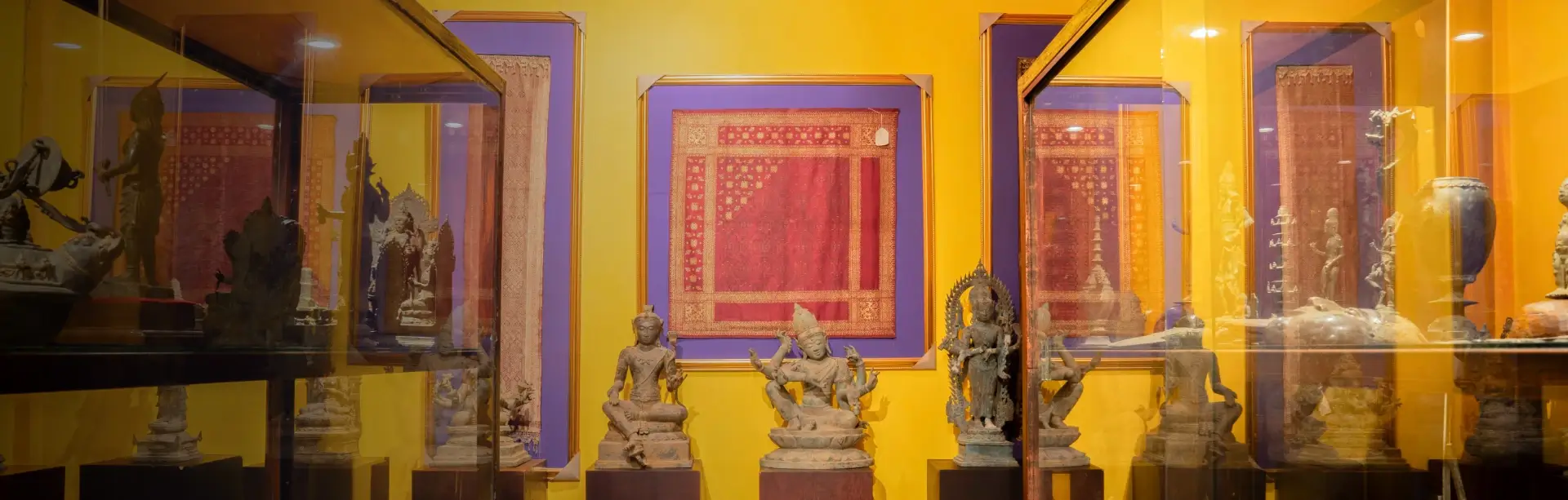 Ancient Hindu-Buddhist bronze statues on dark pedestals with framed traditional textiles, Museum Peter Bloch Collection.